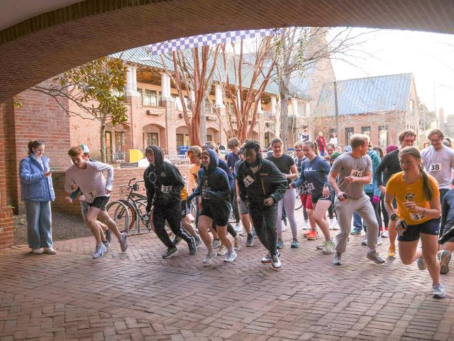 A group of runners at the starting line of a race in an outdoor courtyard, with a few spectators watching nearby.