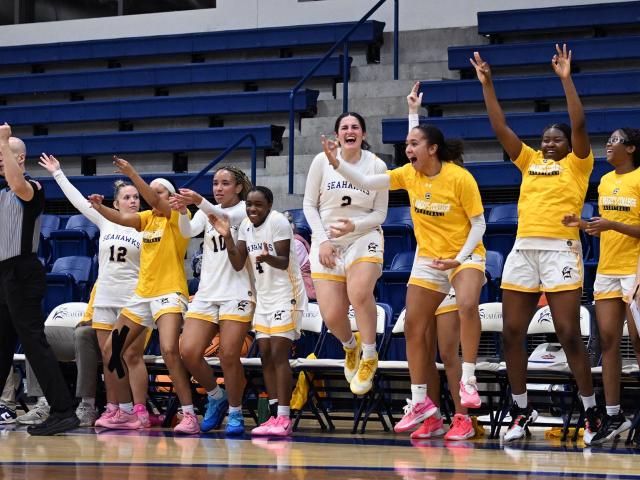 A women's basketball team celebrates enthusiastically on the bench during a game, with players standing, jumping, and raising their arms.