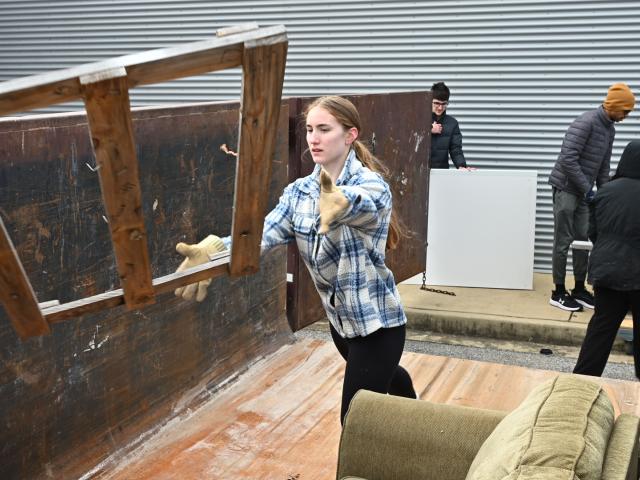 A woman throws a wooden chair into a large dumpster while several other people clear out items nearby.