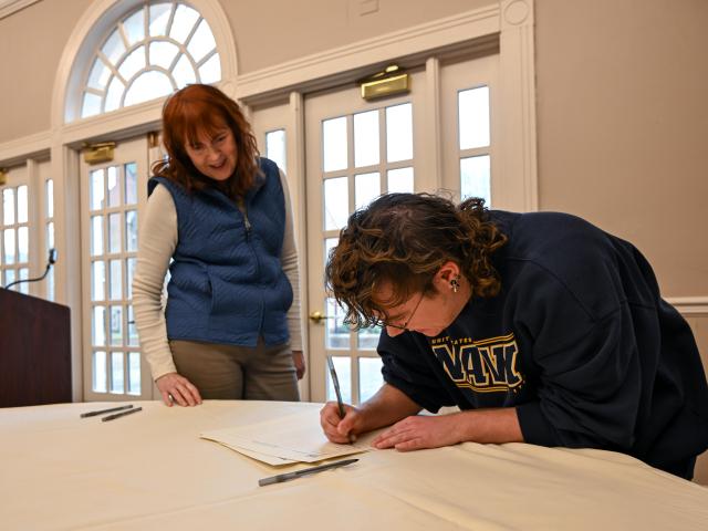A person is bent over a table signing a document while another person stands nearby, watching. Two pens and a microphone are also on the table.