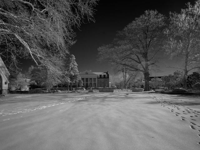 A snow-covered landscape with footprints leading to and from a church building on the left and a large house in the background, surrounded by trees under a clear sky.