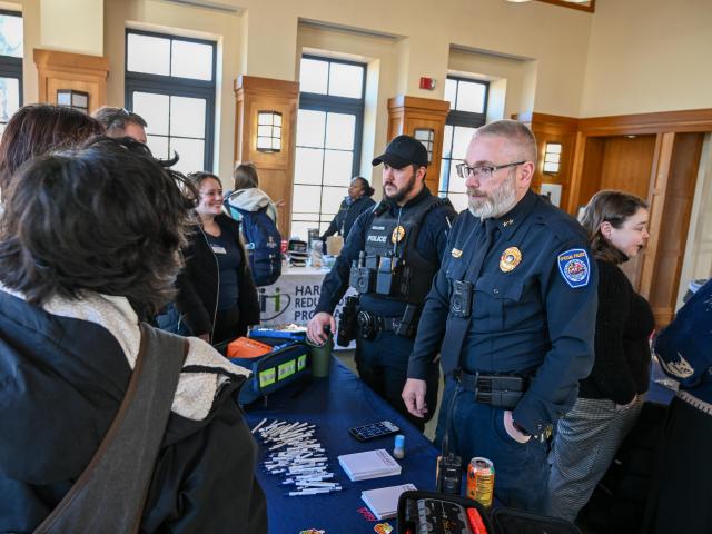 Two uniformed police officers speak to a group of people at an indoor event; informational materials and pens are displayed on the table in front of them.