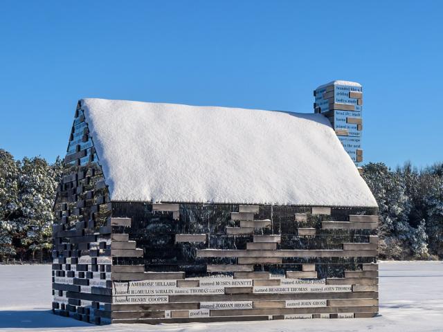 A small wooden cabin covered in snow, with names engraved on its exterior planks, stands in a snowy landscape under a clear blue sky.