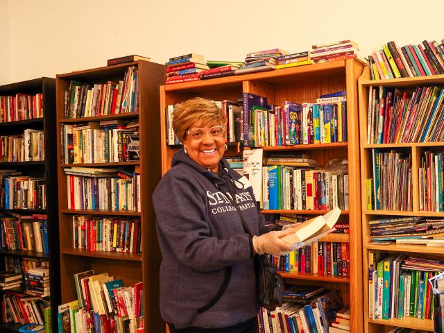 A woman wearing glasses and a hoodie stands in front of bookshelves, holding a book and smiling at the camera in a library or bookstore.