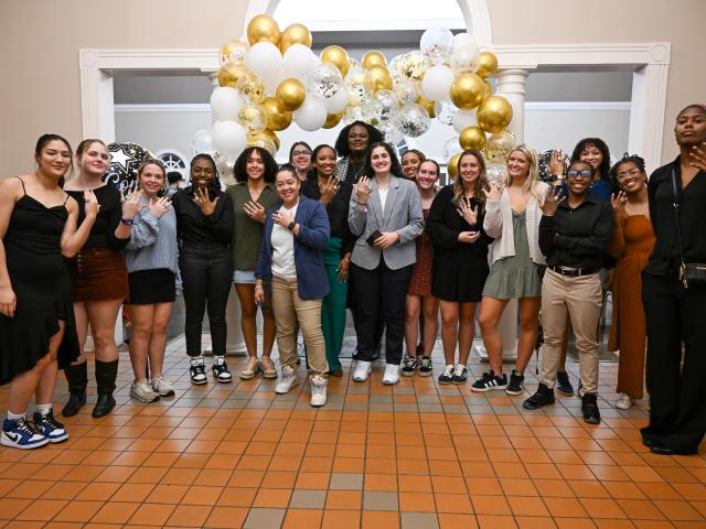 A group of people pose indoors in front of white and gold balloons, many holding up four fingers and smiling at the camera.
