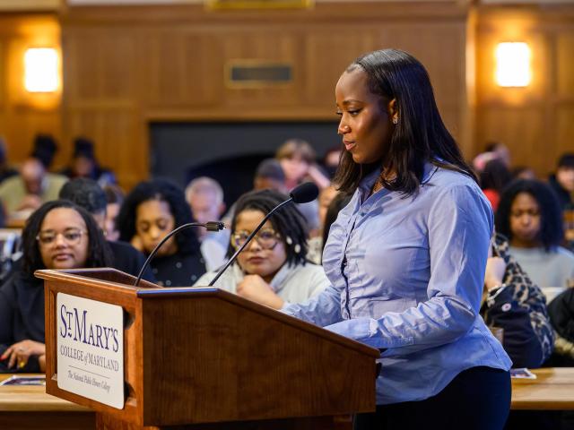 A woman stands at a podium labeled "St. Mary's College of Maryland" speaking to an audience seated in a wood-paneled room.