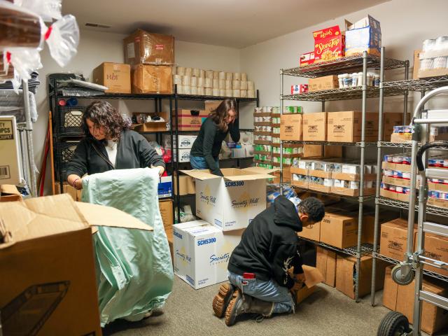Three people organize and pack boxes of food in a storage room filled with shelves stocked with canned goods and other supplies.