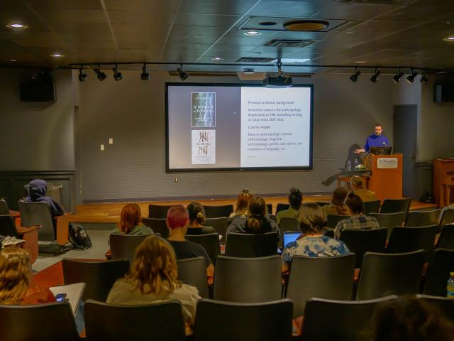 A lecturer stands at a podium presenting slides to an audience seated in an auditorium-style classroom.