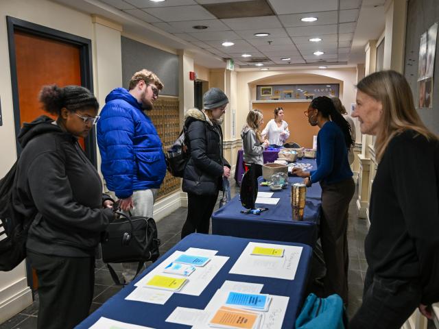 People stand at tables in a hallway, interacting with staff and picking up papers and food items; informational flyers are laid out in the foreground.