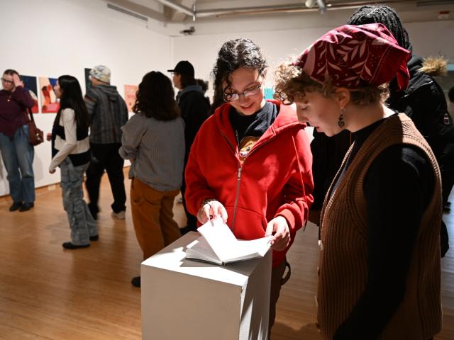 People view art and read a booklet at an indoor gallery exhibition. The room has wooden floors, white walls, and several artworks on display.