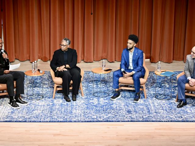 Four people sit on stage in armchairs, engaged in a panel discussion. One person is speaking into a microphone while the others listen. There are small tables with water bottles beside each chair.