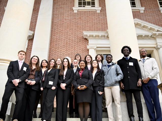 A group of thirteen people in business attire stand together on the steps of a brick building with columns.