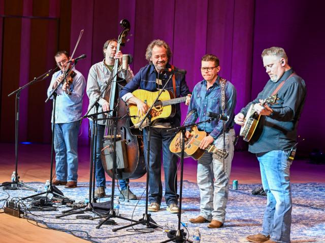 Five musicians perform onstage with fiddle, upright bass, guitar, dobro, and banjo under stage lights, standing on a patterned rug with microphones and cables visible.