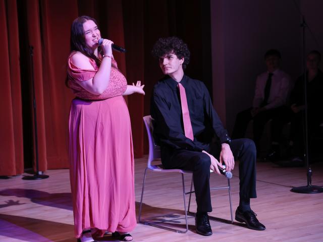 A woman in a pink dress sings into a microphone while standing next to a seated man in a black shirt and pink tie onstage, with red curtains and other people in the background.