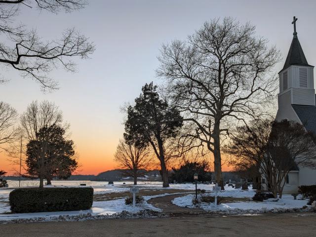 A church with a steeple stands near leafless trees and snow-covered ground at sunset, with a river and distant hills in the background.
