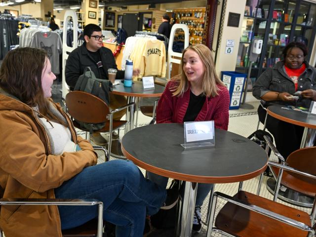 Four students sit at small round tables in a campus store or café. Two women in the foreground talk, while two others use laptops in the background. Shelves of merchandise are visible.