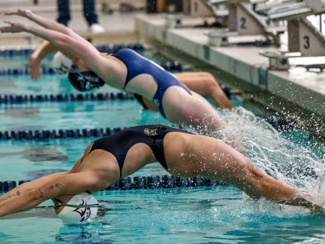 Two competitive swimmers in swimsuits dive backwards into the pool at the start of a backstroke race, with splashes of water visible.