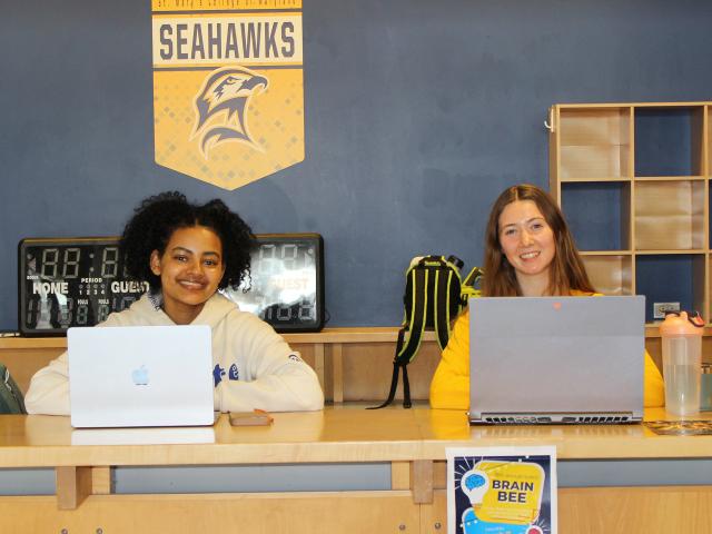 Two women sit at a table with laptops in front of a St. Mary’s College Seahawks sign; a "Brain Bee" event poster is visible on the front of the table.