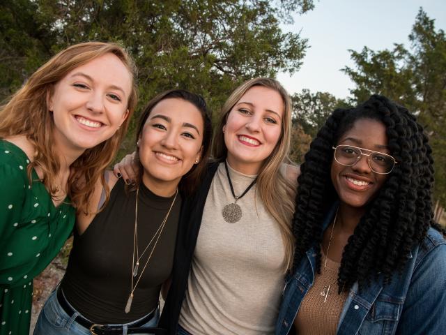 Four women stand close together outdoors, smiling at the camera, with trees and sky in the background.