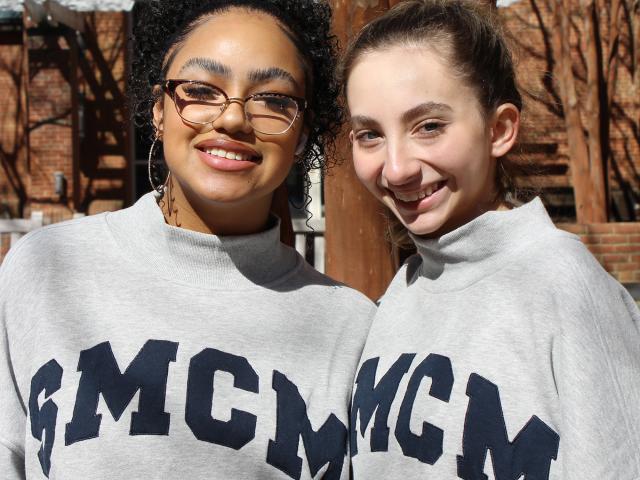 Two young women wearing matching gray sweatshirts with "SMCM" lettering stand outdoors, smiling at the camera.