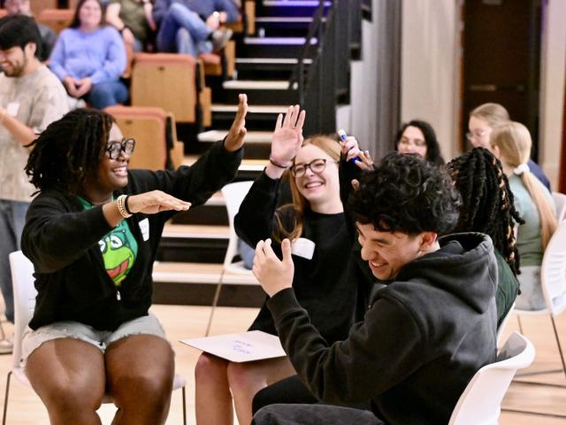 Four young adults sit in a circle indoors, smiling and giving high fives, with others seated in the background.