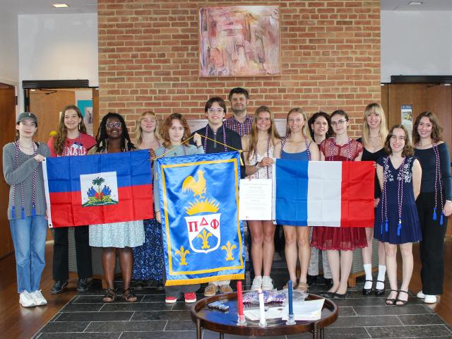 A group of people stands indoors, holding Haitian, French, and society flags, with documents and candles visible on a table in front of them.