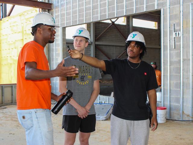 Three people wearing hard hats stand inside a building under construction; one gestures while another holds a recorder toward him.