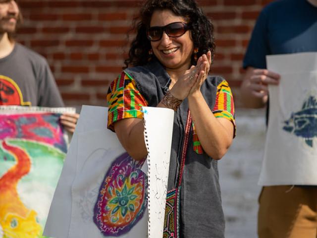 A woman stands outdoors, smiling and clapping, holding colorful artwork. She is flanked by two people also holding art, with a brick wall in the background.