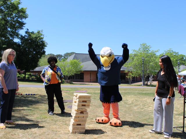 Four people, including one in an eagle mascot costume, stand around a giant Jenga tower outdoors on a sunny day. The mascot has its arms raised in celebration.