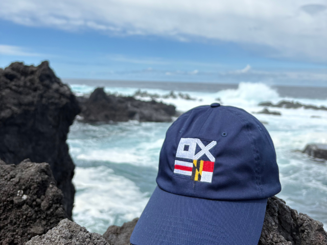 A blue baseball cap with multicolored flag designs rests on dark volcanic rocks by the ocean, with waves and a cloudy sky in the background.