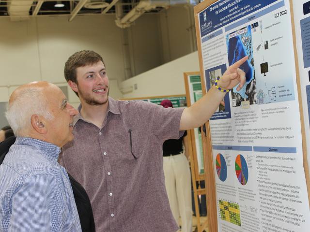 Two men stand in front of a scientific poster display; the younger man points at the poster while explaining information to the older man.