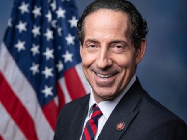 A man in a dark suit and striped tie stands in front of a U.S. flag, smiling at the camera against a blue background.