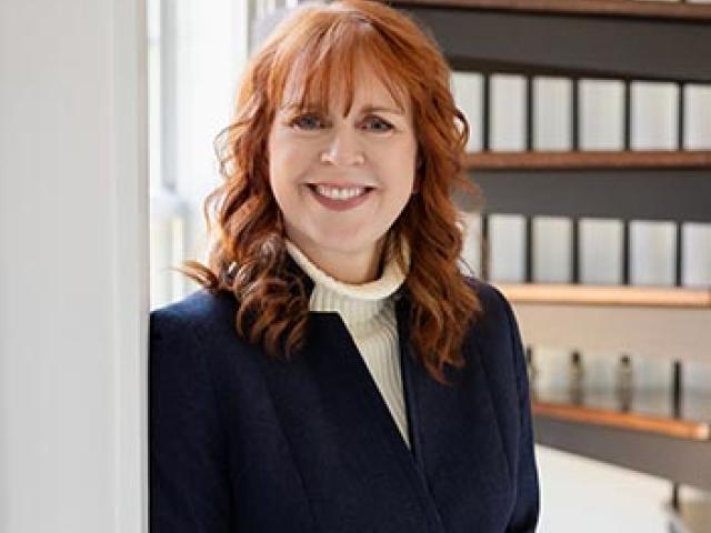 A woman with red hair, wearing a dark blazer and light turtleneck, stands indoors near a window, smiling with hands clasped. A spiral staircase is visible in the background.