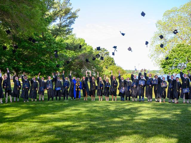 A group of graduates in caps and gowns stand on a lawn, tossing their caps into the air in celebration, surrounded by green trees under a clear sky.