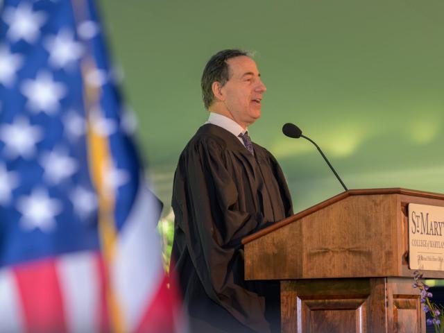 A man in academic robes speaks at a podium labeled "St. Mary’s College" during an outdoor event, with an American flag visible in the foreground.