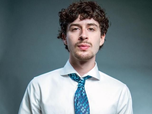 A young man with curly hair wearing a white shirt and blue patterned tie stands against a plain gray background, looking directly at the camera.