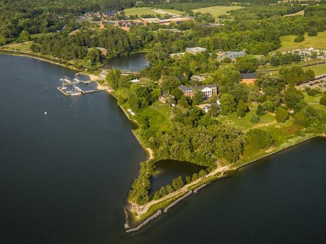Aerial view of a green, tree-covered peninsula with buildings, docks, and boats along the shoreline, bordered by a river or lake.