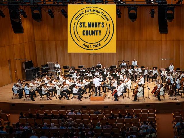 A symphony orchestra performs on stage in a wooden concert hall with a banner overhead reading "Music for Maryland, St. Mary's County, Aug. 1, 2025." Audience members are seated below.