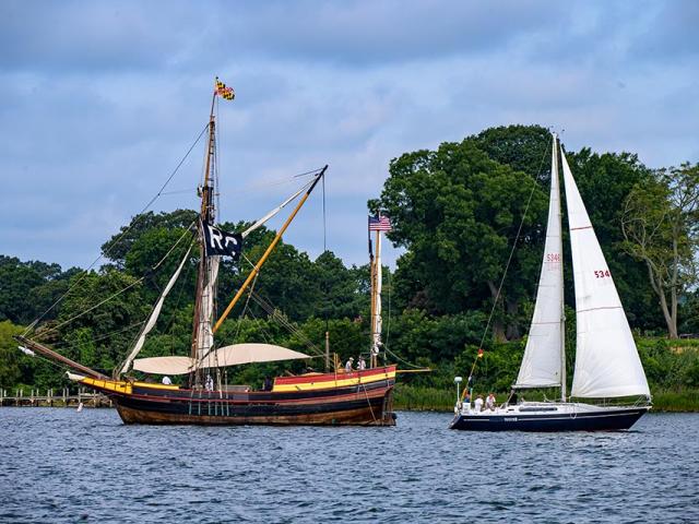 A wooden tall ship with a race committee flag and a modern white sailboat are side by side on a river, with trees and greenery in the background.