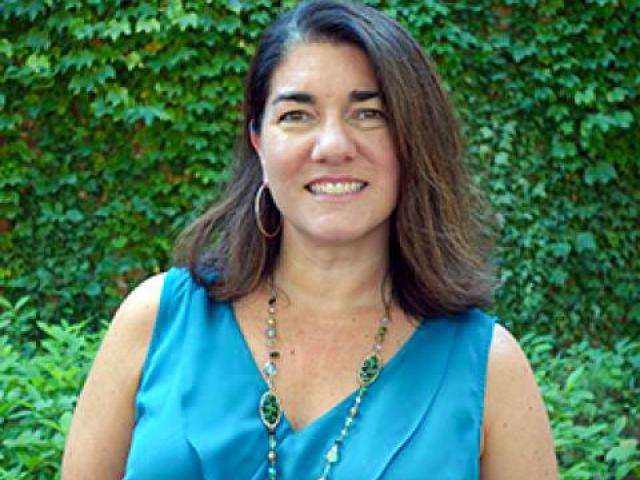 A woman with shoulder-length brown hair, wearing a blue sleeveless top and a beaded necklace, stands outdoors in front of a leafy green wall.