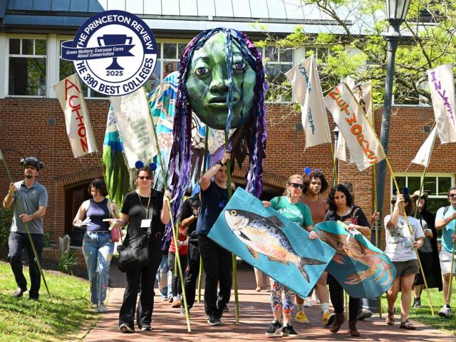A group of people participate in an outdoor parade carrying banners, a large fish prop, and a green puppet head. A "Best 390 Colleges" Princeton Review badge is overlaid in the top left.