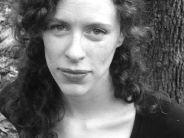 A woman with curly hair and a serious expression looks at the camera. She is wearing a dark top and standing outdoors near a tree. The image is in black and white.