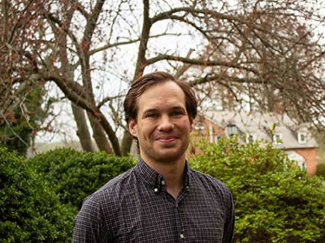 A man stands outdoors in front of green bushes and leafless trees, wearing a checked button-up shirt, smiling at the camera.