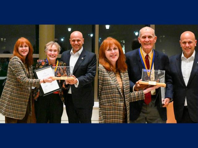 Six people stand indoors, with two women and one man holding awards and certificates, all dressed in formal or business attire, smiling at the camera.