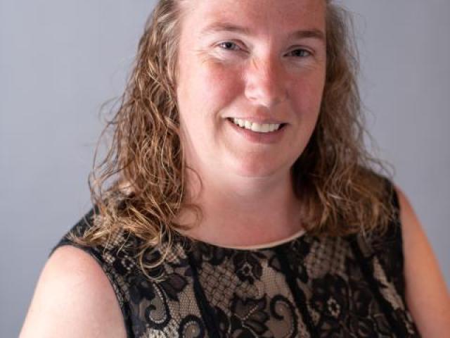 Woman with wavy, shoulder-length hair wearing a black lace dress, smiling, and posed against a plain gray background.