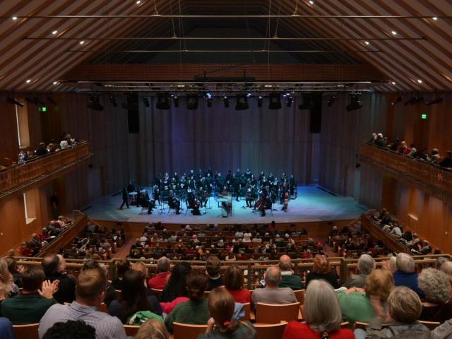 A symphony orchestra performs on stage in a wooden concert hall, with audience members seated on the main floor and balconies.