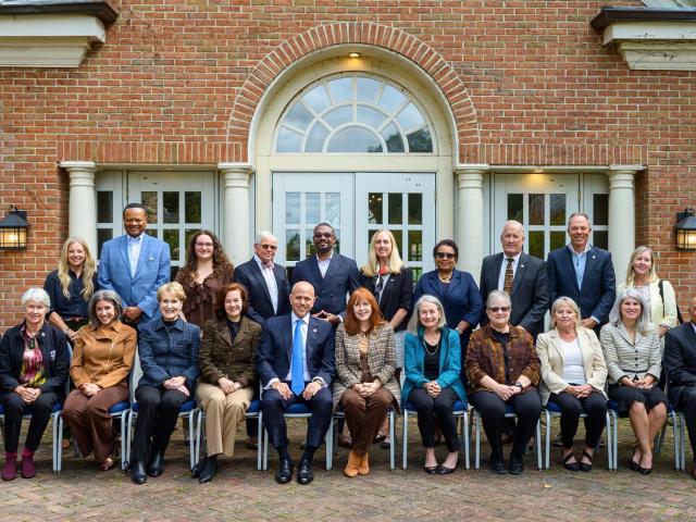 A group of 24 adults pose for a formal group photo in two rows outside a brick building with arched windows and columns.