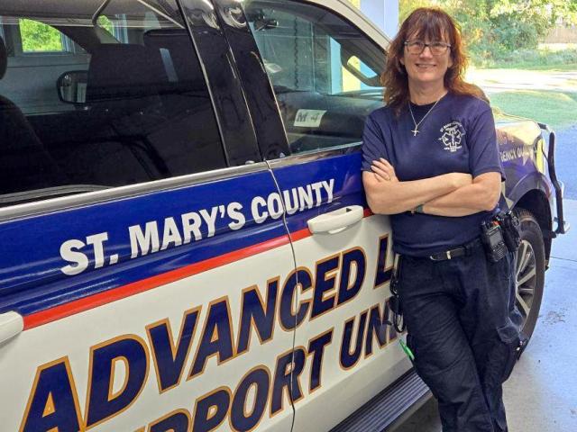 A person in a navy uniform stands smiling and leaning against a St. Mary's County Advanced Life Support Unit vehicle, parked inside a garage.