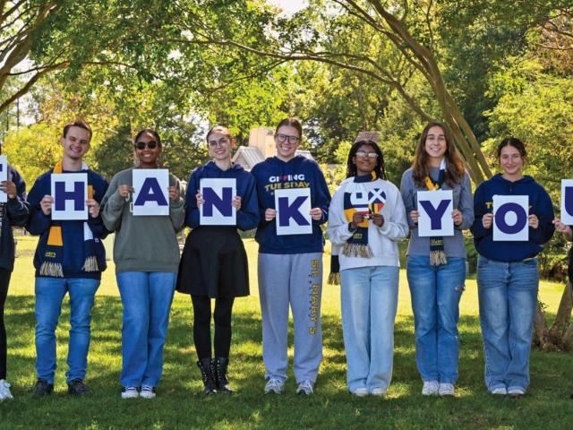 Eight people stand outdoors in a row, each holding a letter sign that together spell out "THANK YOU," with trees and greenery in the background.