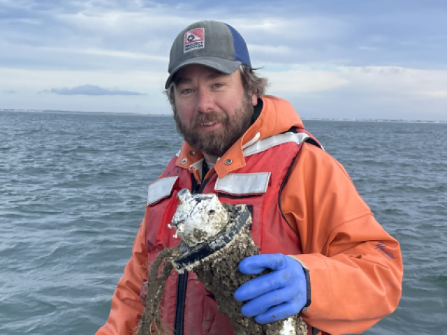 A man in an orange life jacket and blue gloves stands on a boat holding a barnacle-covered object over the water under a cloudy sky.